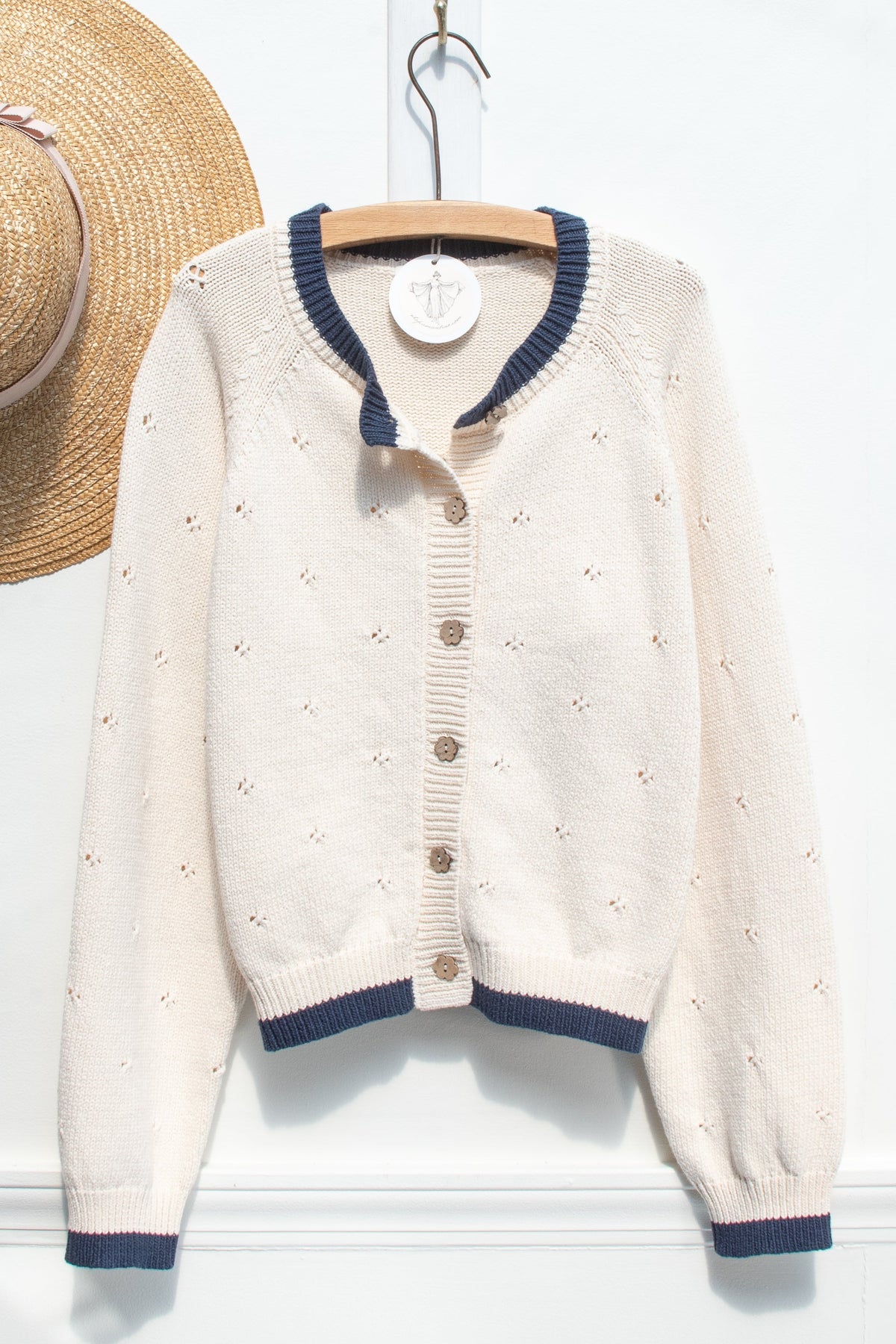 A cream-colored knit cardigan sweater with navy blue trim, hanging on a wooden hanger against a white wall. A straw hat is partially visible on the left side of the frame.