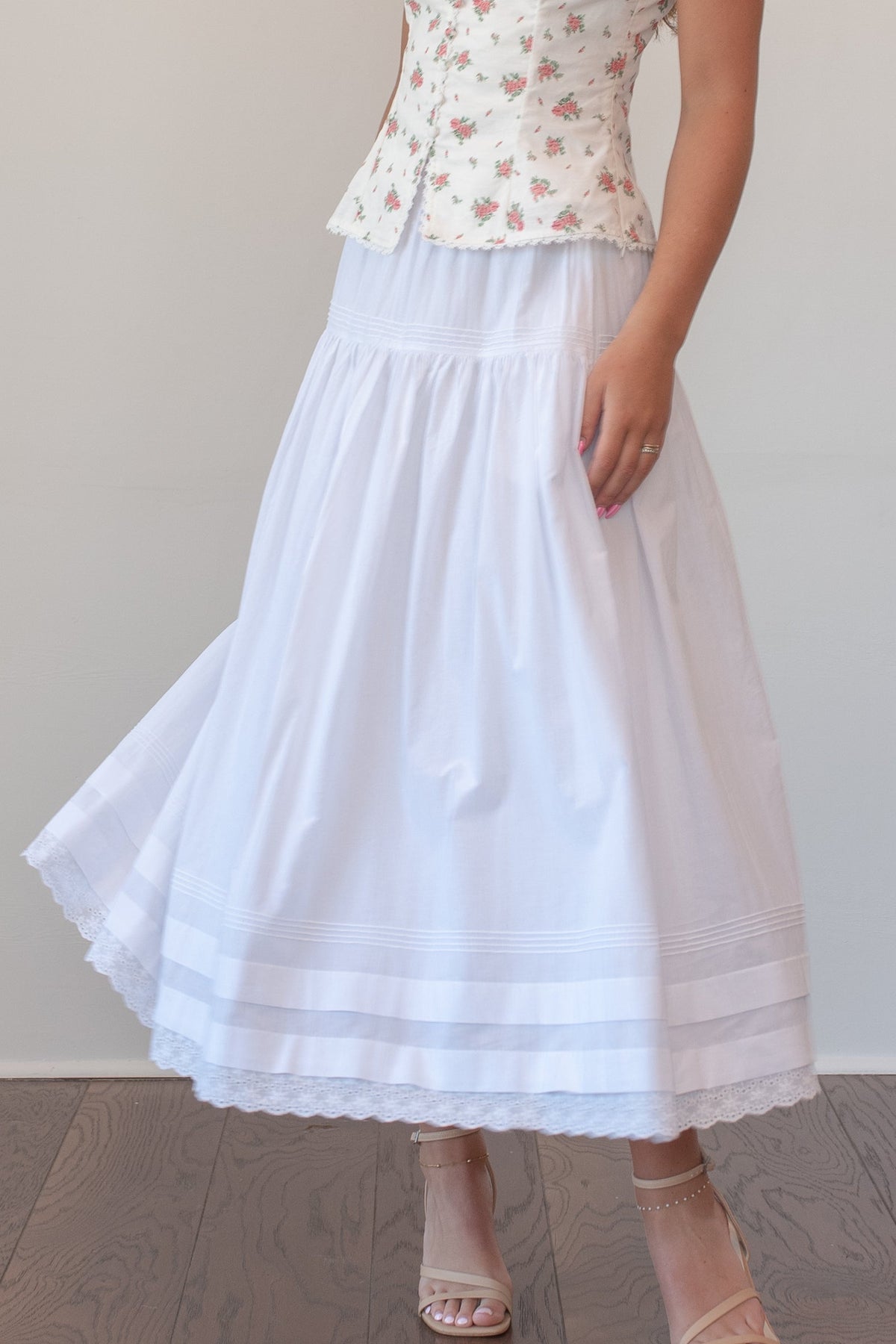 A woman stands indoors wearing a sleeveless white corset-style top with a small pink floral print and a long white skirt, paired with nude high-heel sandals. The background is a plain white wall with dark wood flooring.