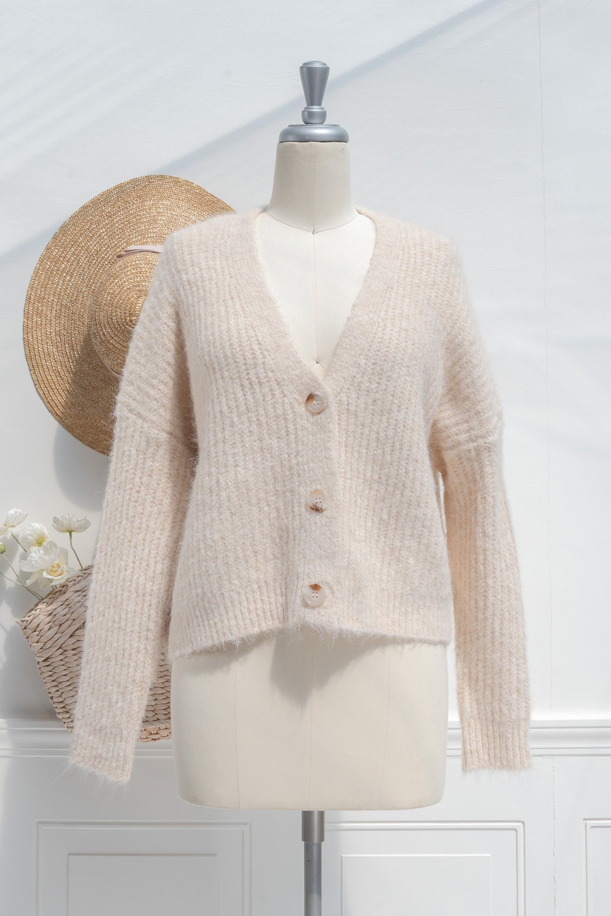 Beige knitted cardigan on a mannequin with a straw hat and flowers in the background.
