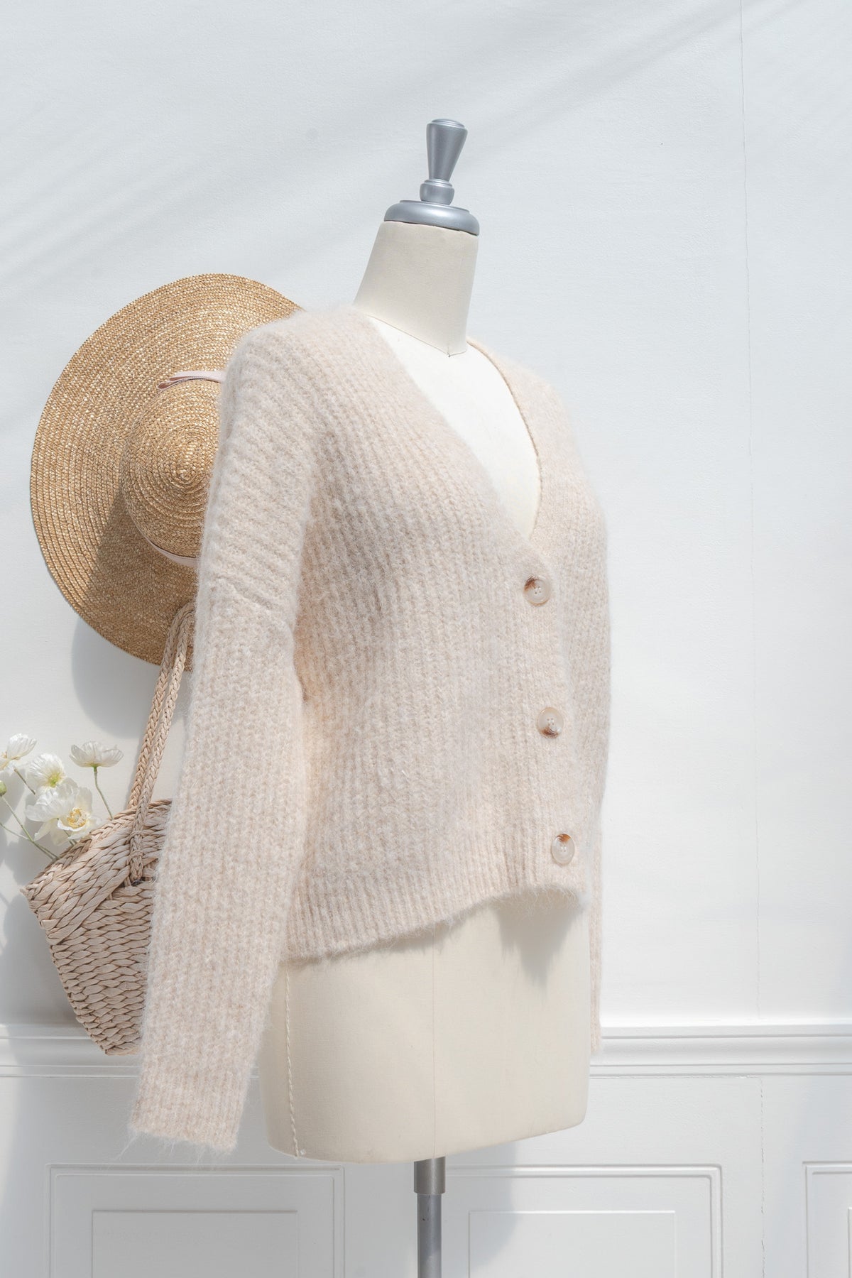 Mannequin wearing a beige knitted cardigan with a straw hat and basket against a white wall.