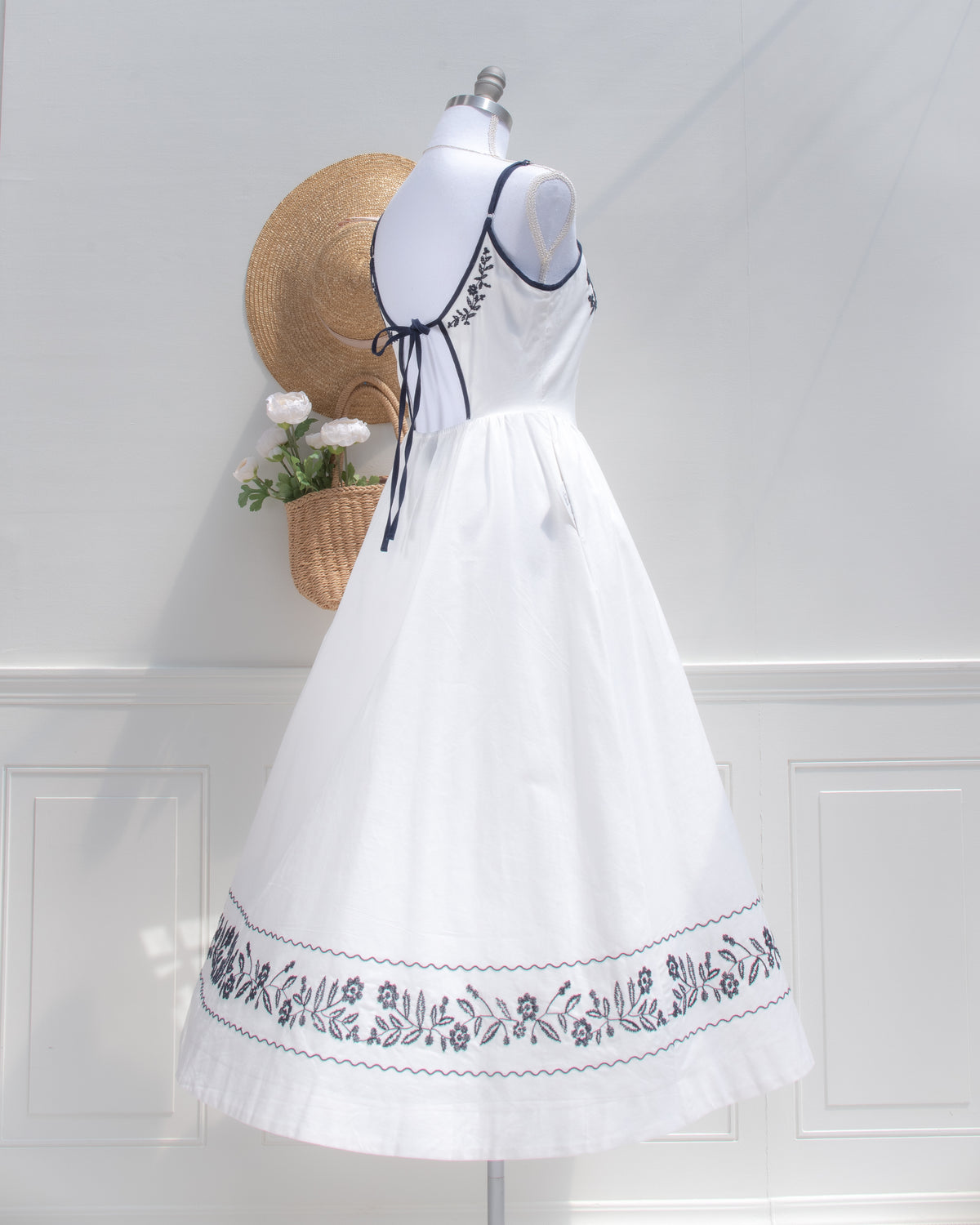 Three-quarter back view of the white embroidered dress, showing the navy tie, full skirt and embroidered hem border.