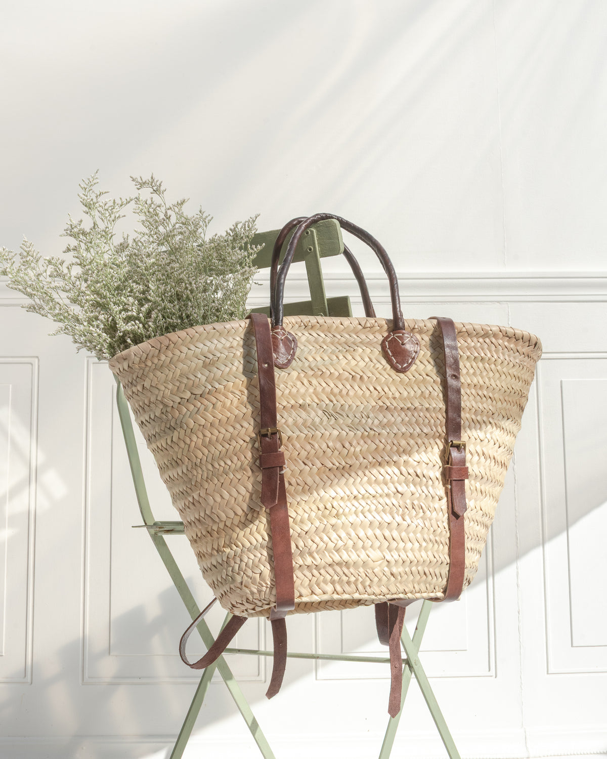 Woven straw tote set on a green bistro chair, handles upright and straps buckled at the sides.
