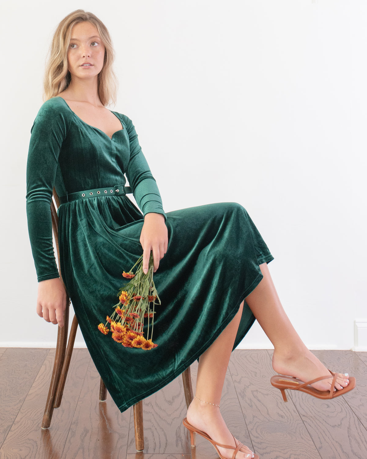 Model seated on a wooden chair, showing the long sleeves, belted waist and full skirt of the emerald velvet dress.