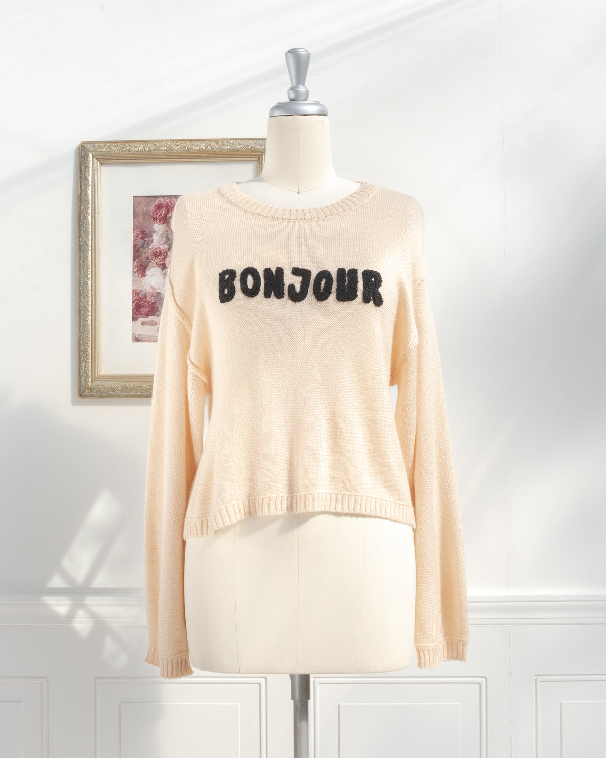 Beige knit sweater with raised black Bonjour appliqué on a mannequin, French girl style, front view.