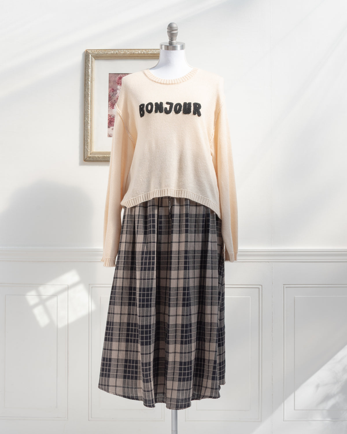 Beige knit sweater with Bonjour appliqué styled with a plaid midi skirt on a mannequin, French girl style, front view.