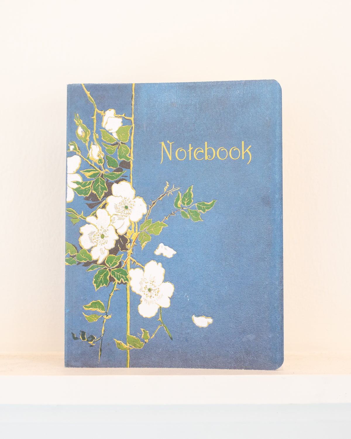 Blue vintage notebook with a white rose cover and gold lettering resting on a white shelf, front view.