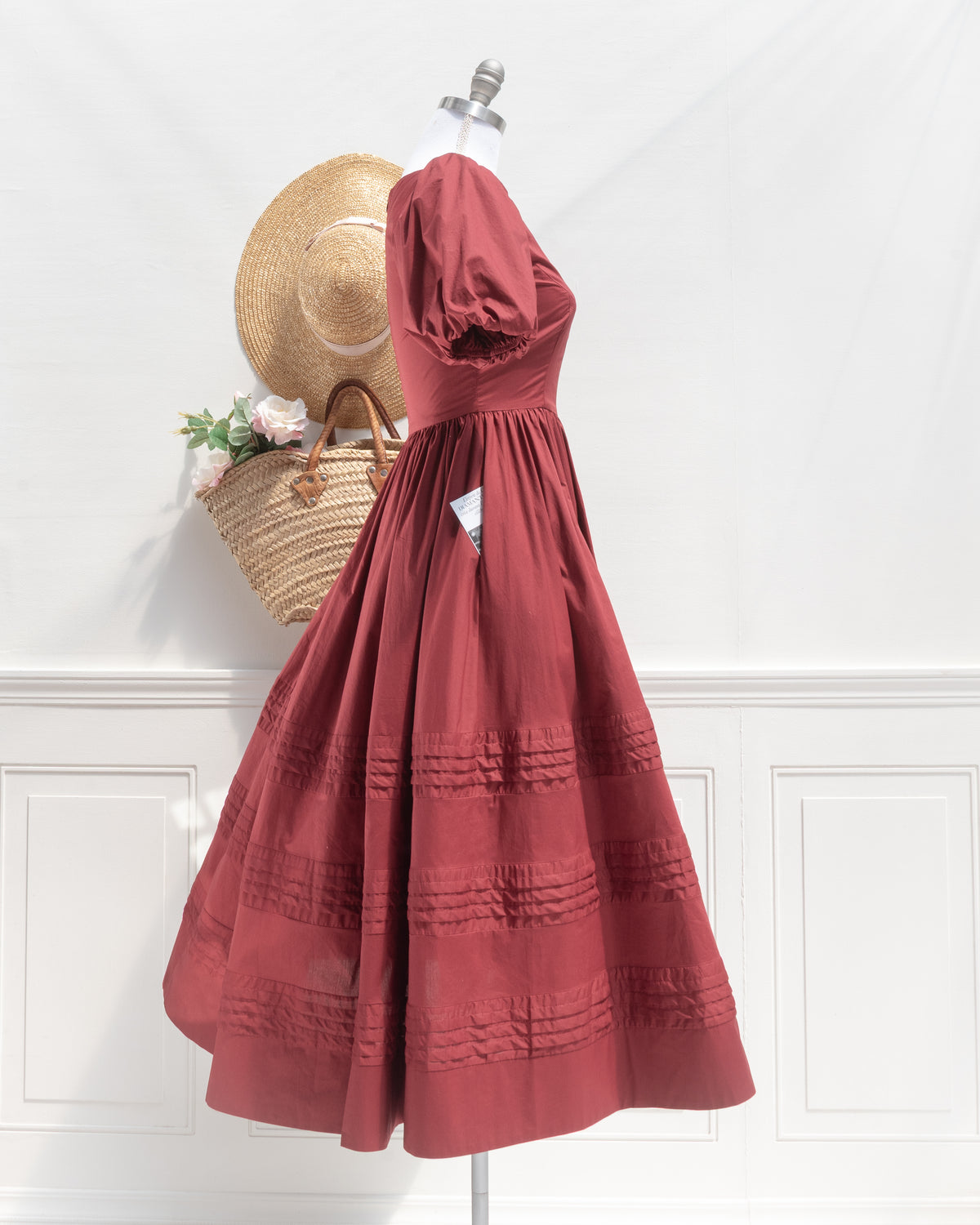 Side profile of the Felicity Dress in burgundy cotton on a dress form, showing the puff sleeve, fitted bodice, side pocket, and pintucked midi skirt.