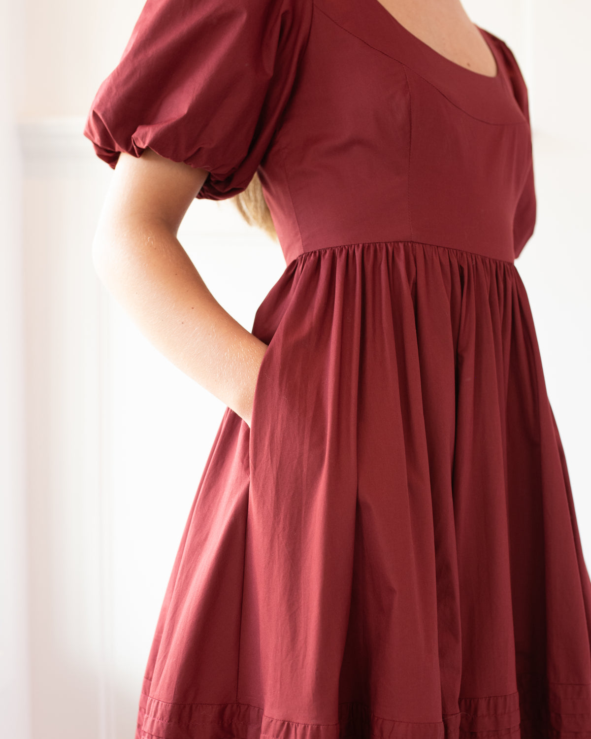Fashion model wearing a burgundy midi dress showing puff sleeves, round neckline, and side pocket detail. Feminine French Girl style dress outfit from Amantine.
