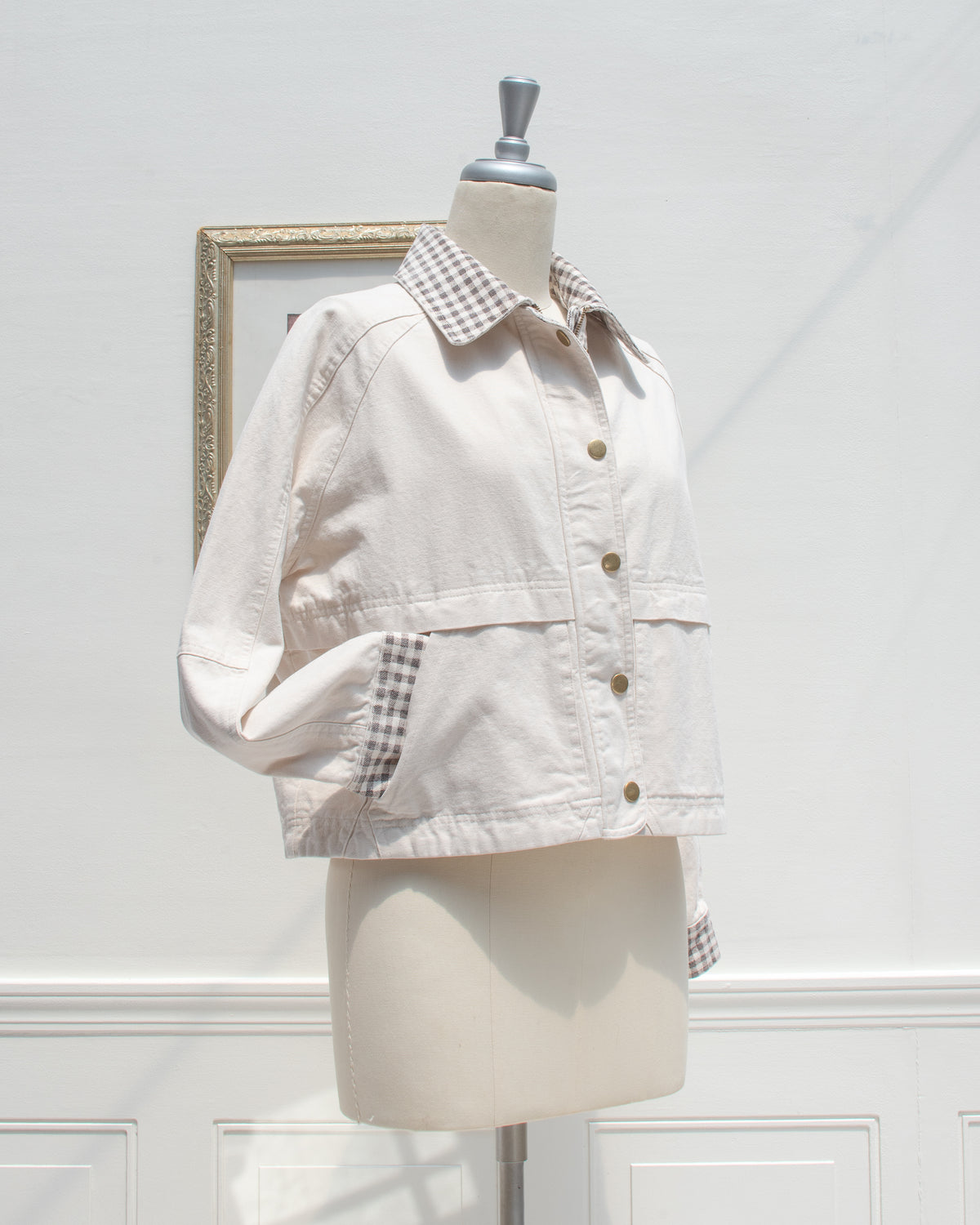 Three-quarter view of the Marlowe Jacket in cream cotton with the gingham collar folded down, brass snap front, and a sleeve tucked into the side pocket.