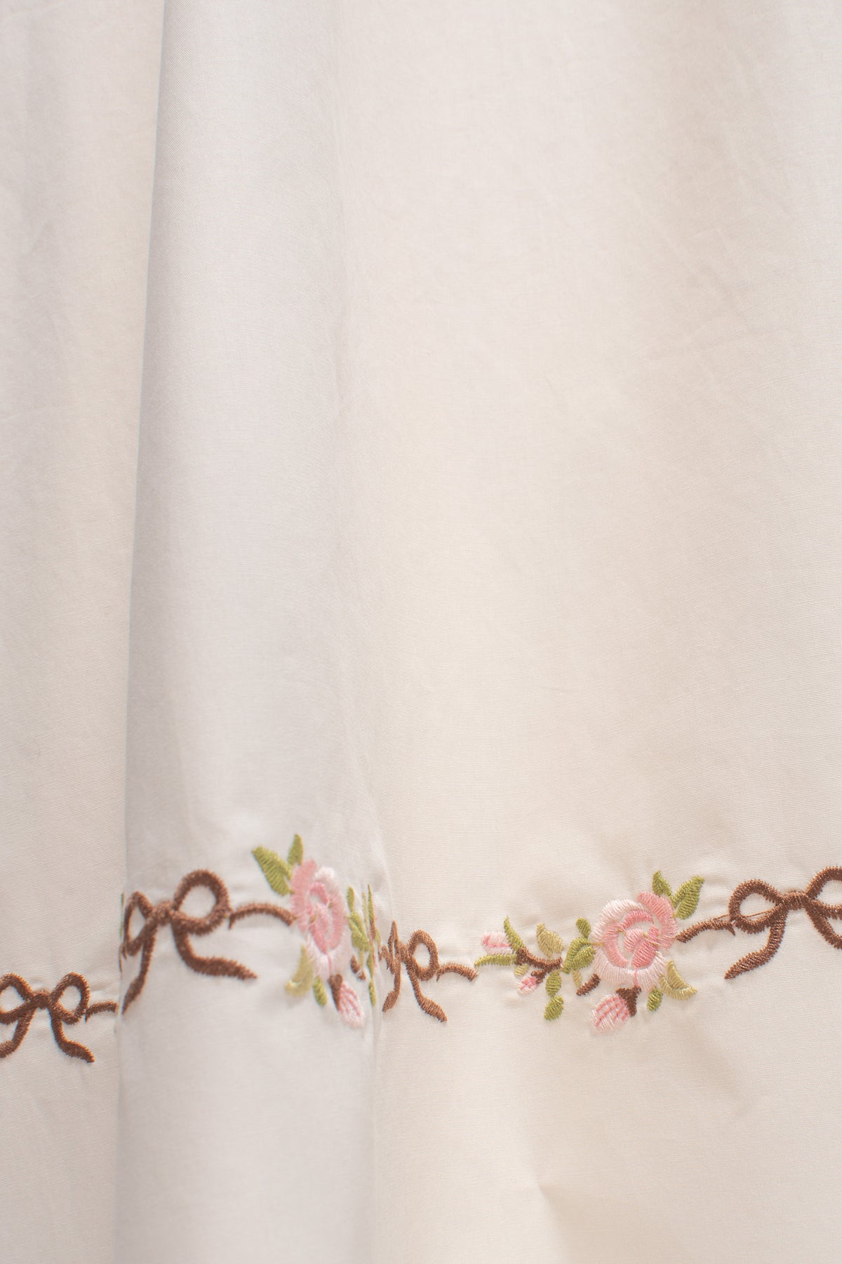 Cream cotton fabric with pink rose and brown bow embroidered motif hem close up.