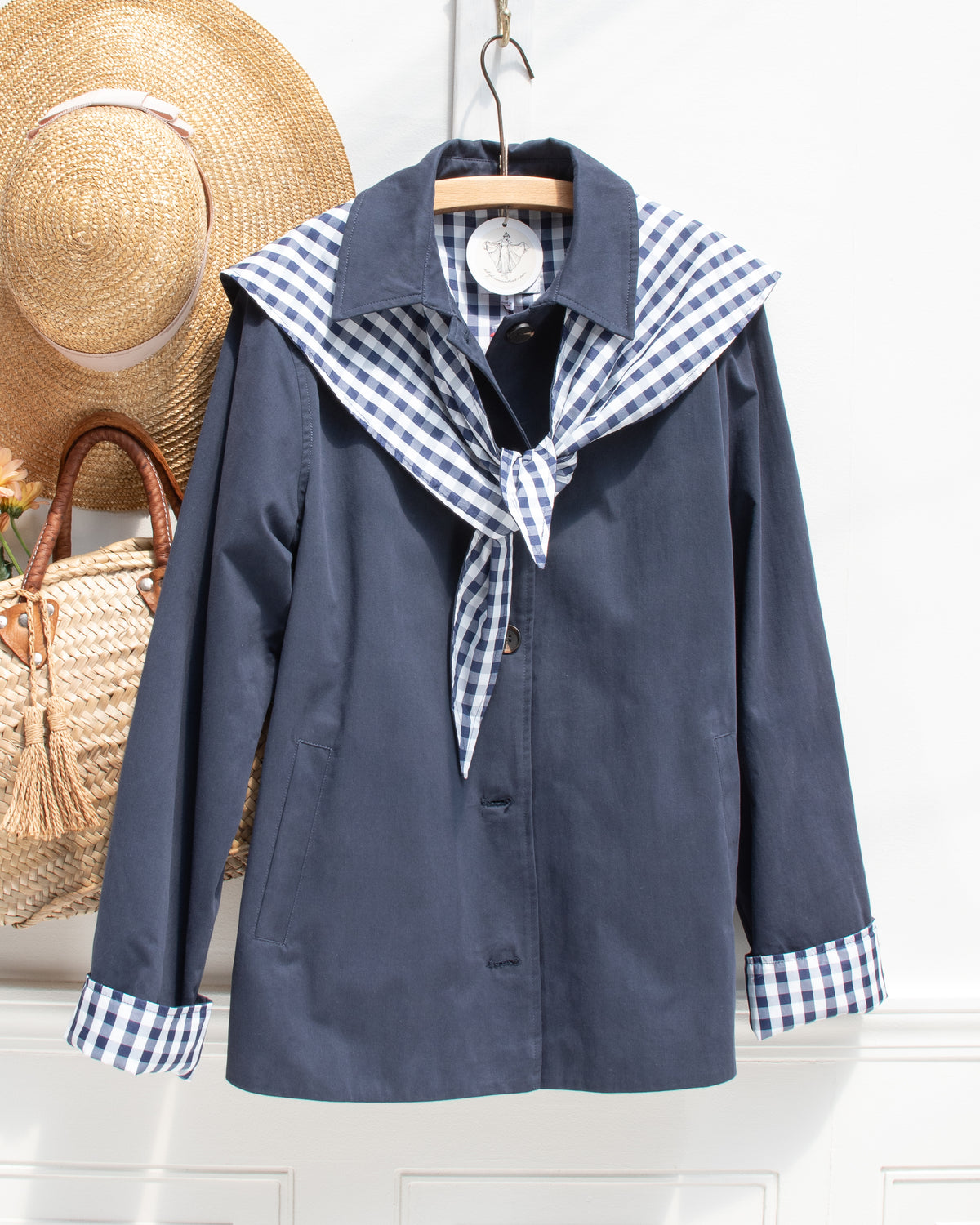 Romantic and feminine clothing, French girl style outerwear. A navy jacket with folded gingham cuffs shown on a hanger, front view. Amantine.