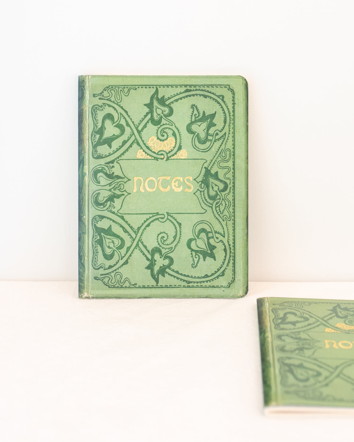 Two green vintage notebooks with ornate vine covers and gold lettering, one upright and one in the foreground, front view.
