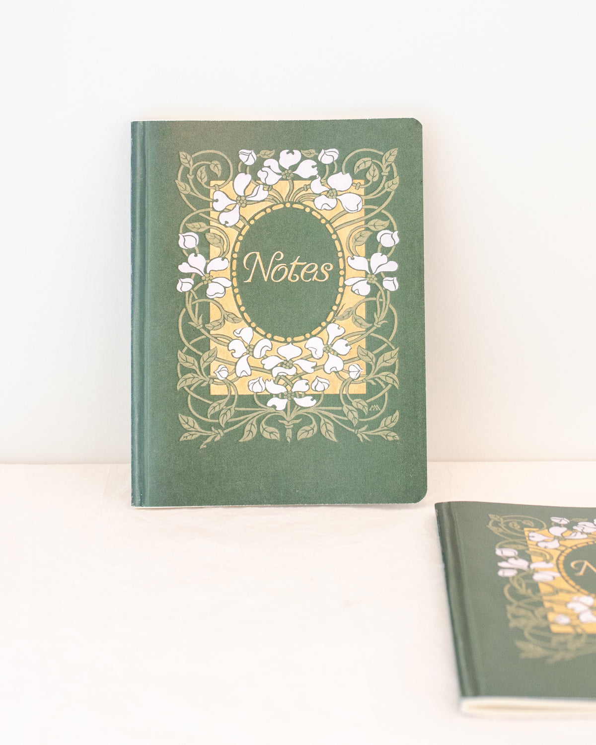Green vintage notebook with white dogwood blossoms and a gold panel, one upright and one in the foreground, front view.