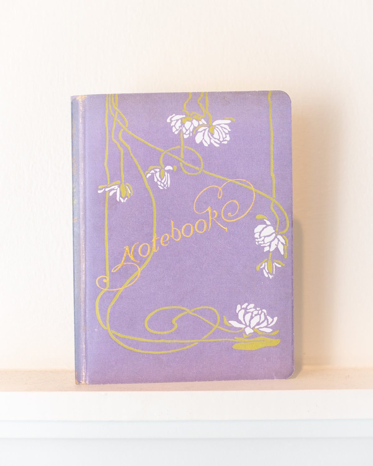 Lavender vintage notebook with a white blossom cover and gold script lettering resting on a white shelf, front view.