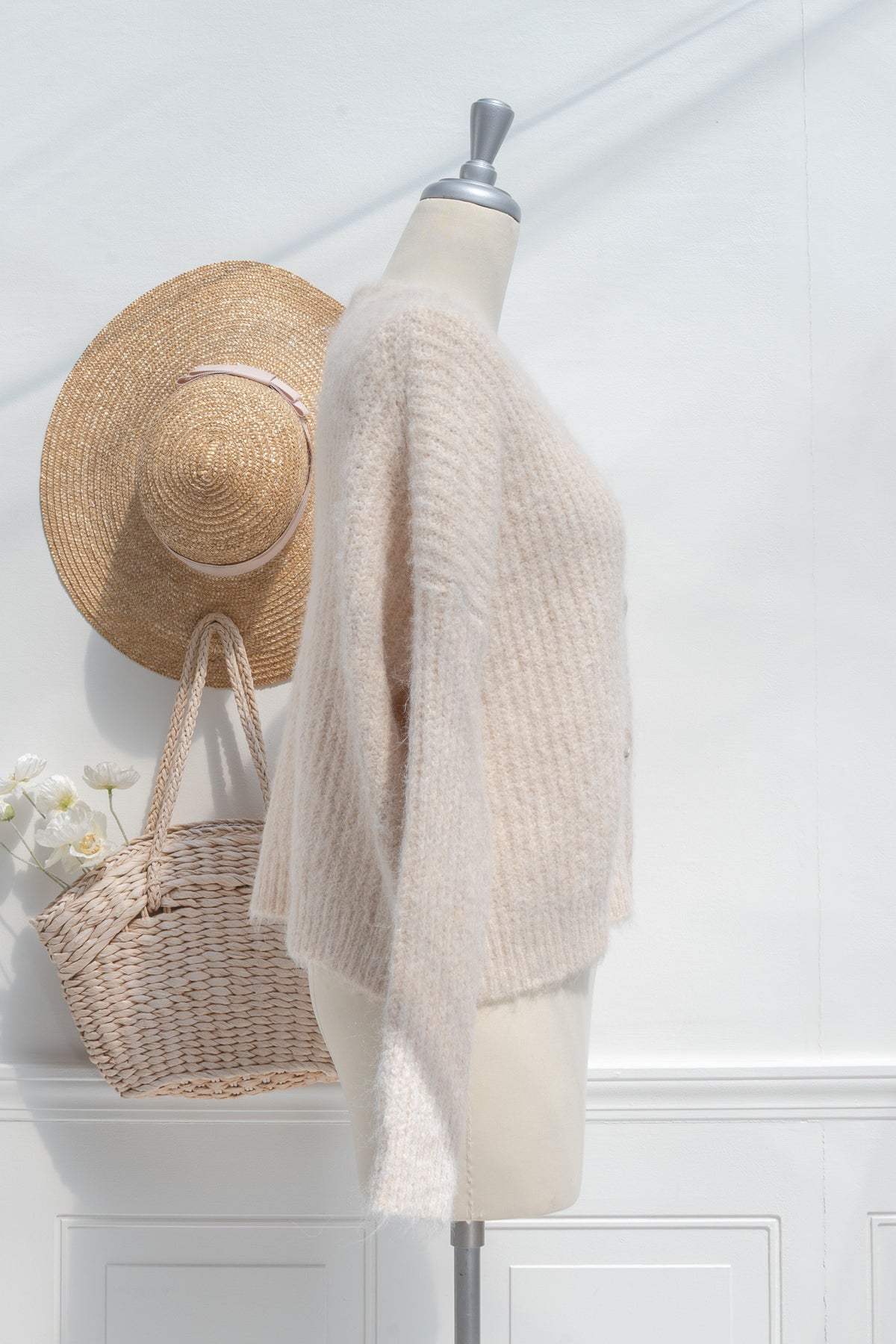 Mannequin wearing a beige knitted cardigan with a straw hat and basket against a white wall.