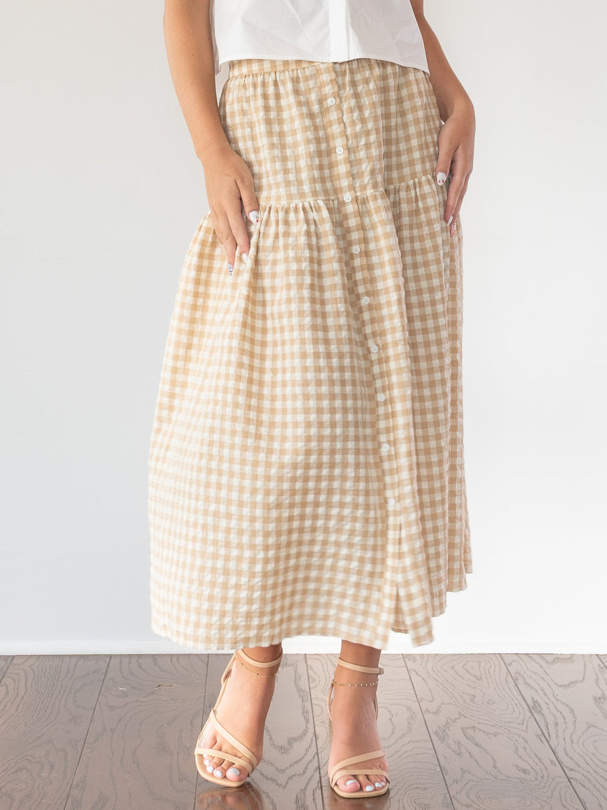 A woman modeling a white short-sleeve button-up shirt with a decorative collar and a beige and white gingham tiered maxi skirt, standing in a bright room with a white wall and wooden floor.