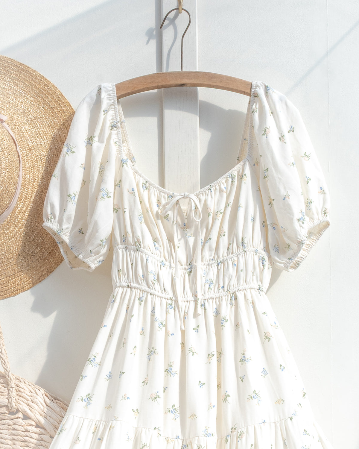 On a hanger, the cream floral dress shows a front neckline tie, gathered bodice and smocked elastic waist.
