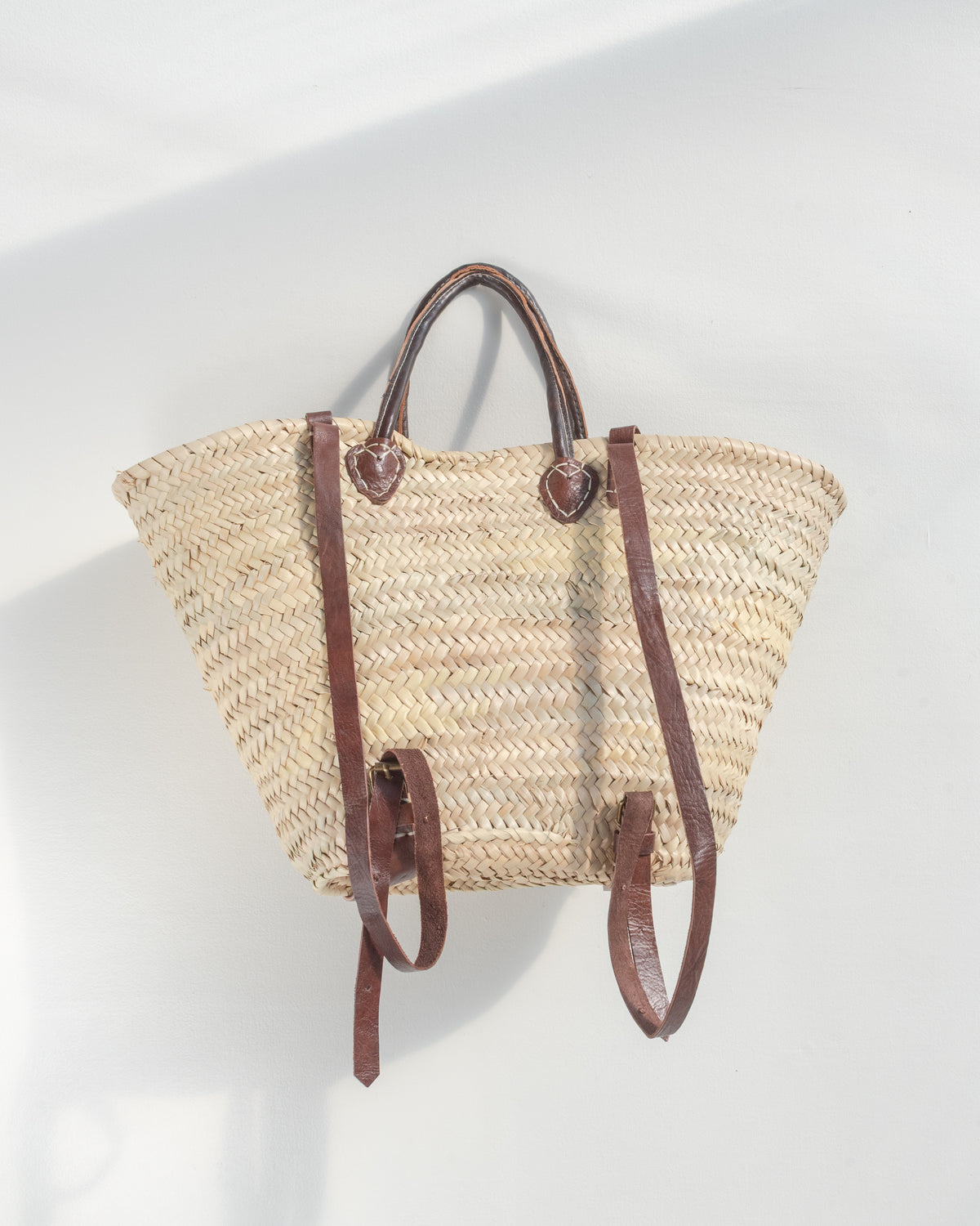 Back view of the straw tote worn as a backpack, two adjustable brown straps with brass buckles.