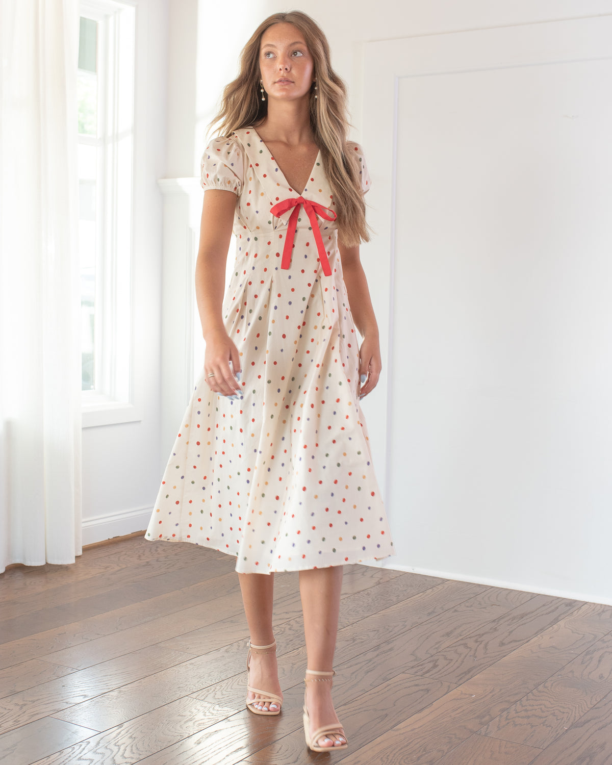 Worn by a model with nude strappy sandals, the cream polka dot dress has a V neckline and graceful midi skirt.