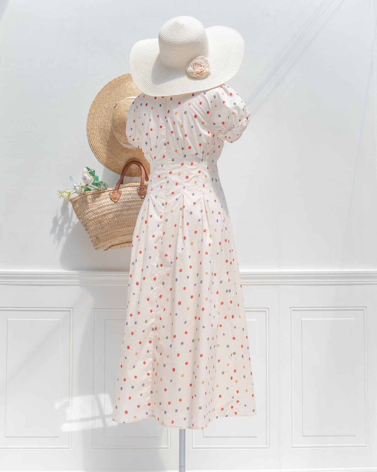 Styled with a white straw sun hat trimmed with a fabric rose, the cream polka dot dress seen from the back.