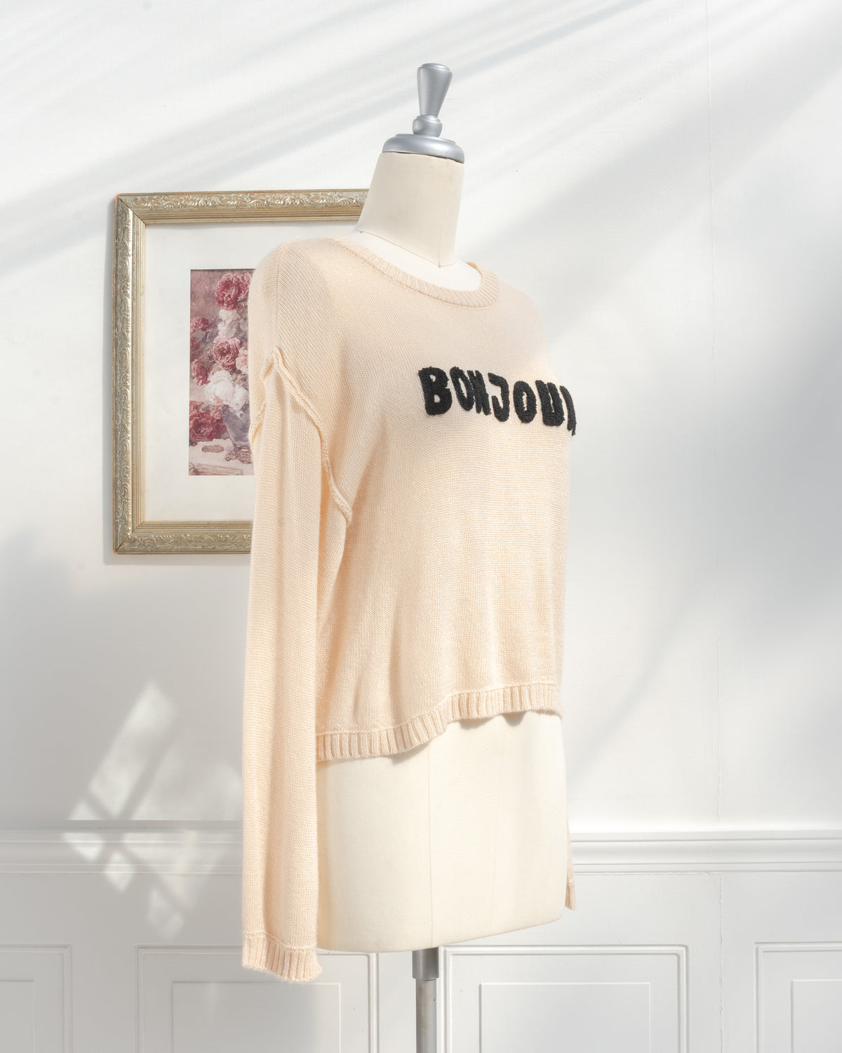 Beige knit sweater with Bonjour appliqué and high low hem on a mannequin, classic fall season, three quarter view.