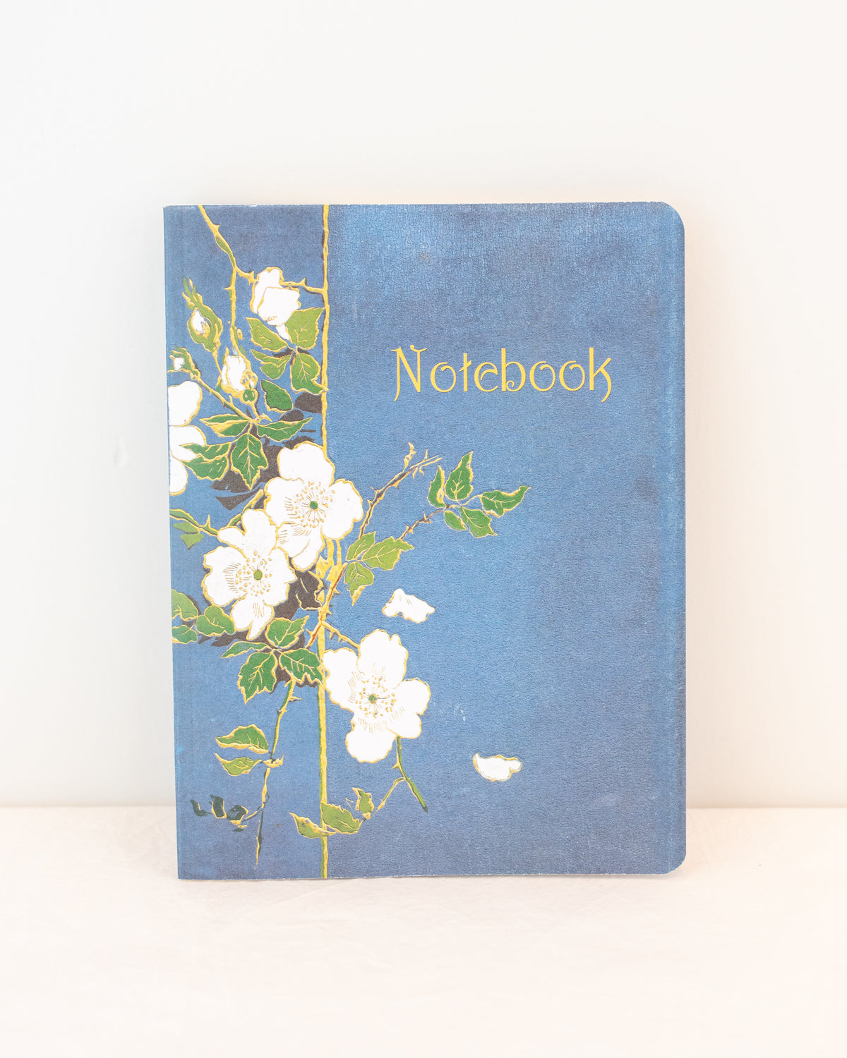 Blue vintage notebook with a white rose cover and gold lettering that reads Notebook, styled upright, front view.