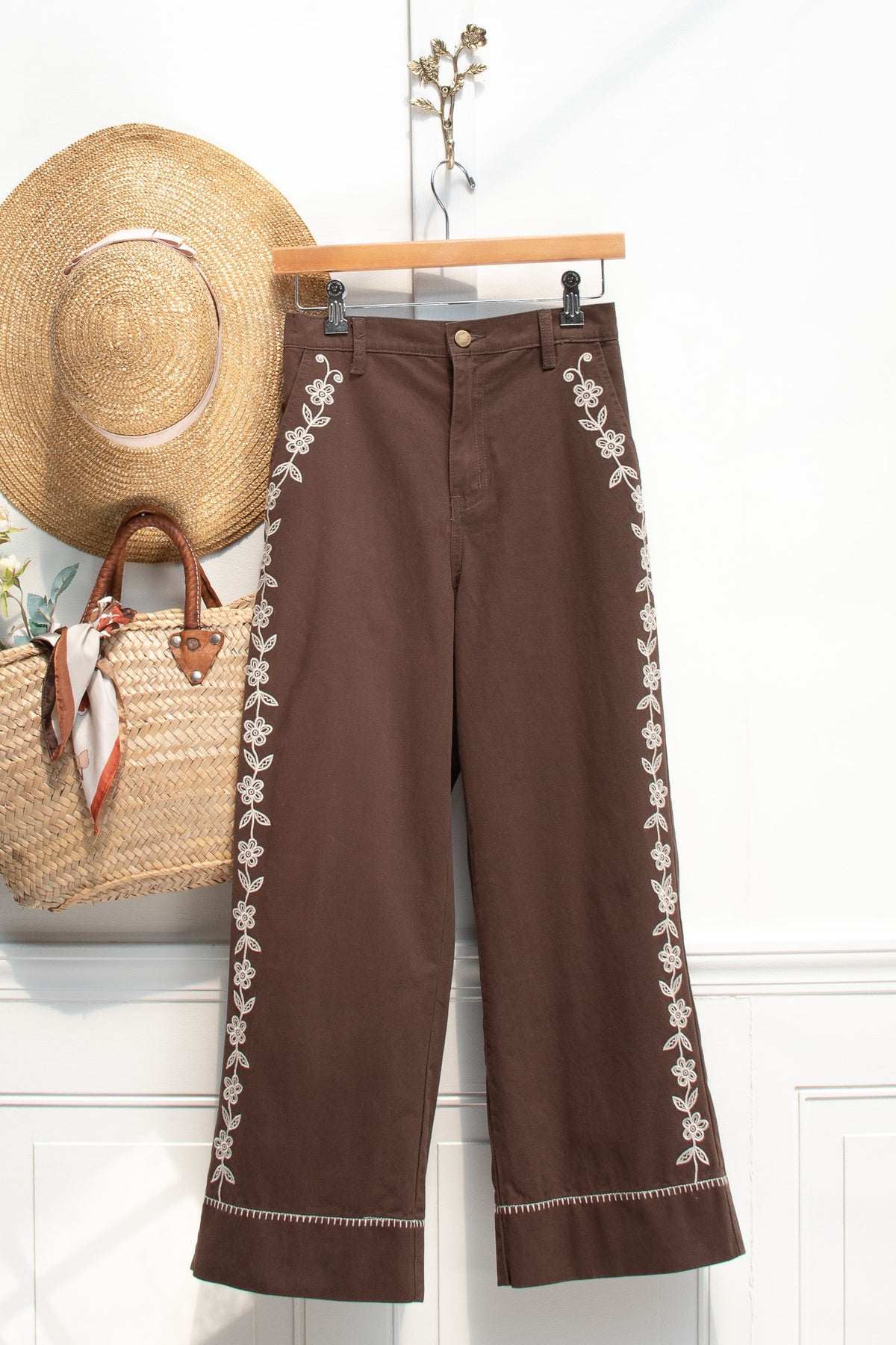 Brown cotton denim pants with floral embroidery, a wardrobe staple displayed on a hanger, front view.