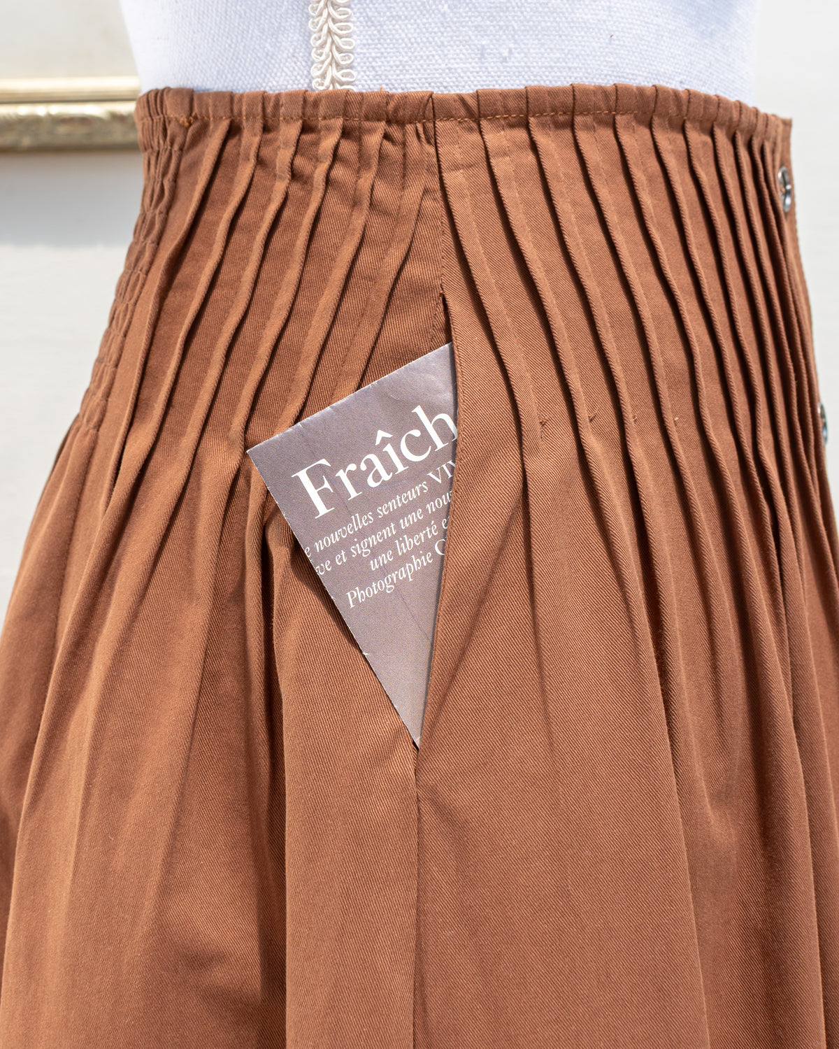 Brown cotton midi skirt with pleated waist and hidden side pocket, close mannequin view.