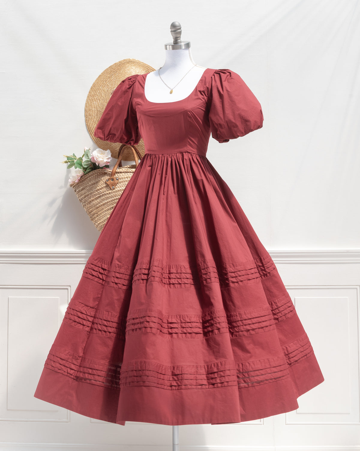 Felicity Dress in burgundy cotton on a dress form with a straw hat and market basket of roses, a French romantic style puff sleeve midi dress.