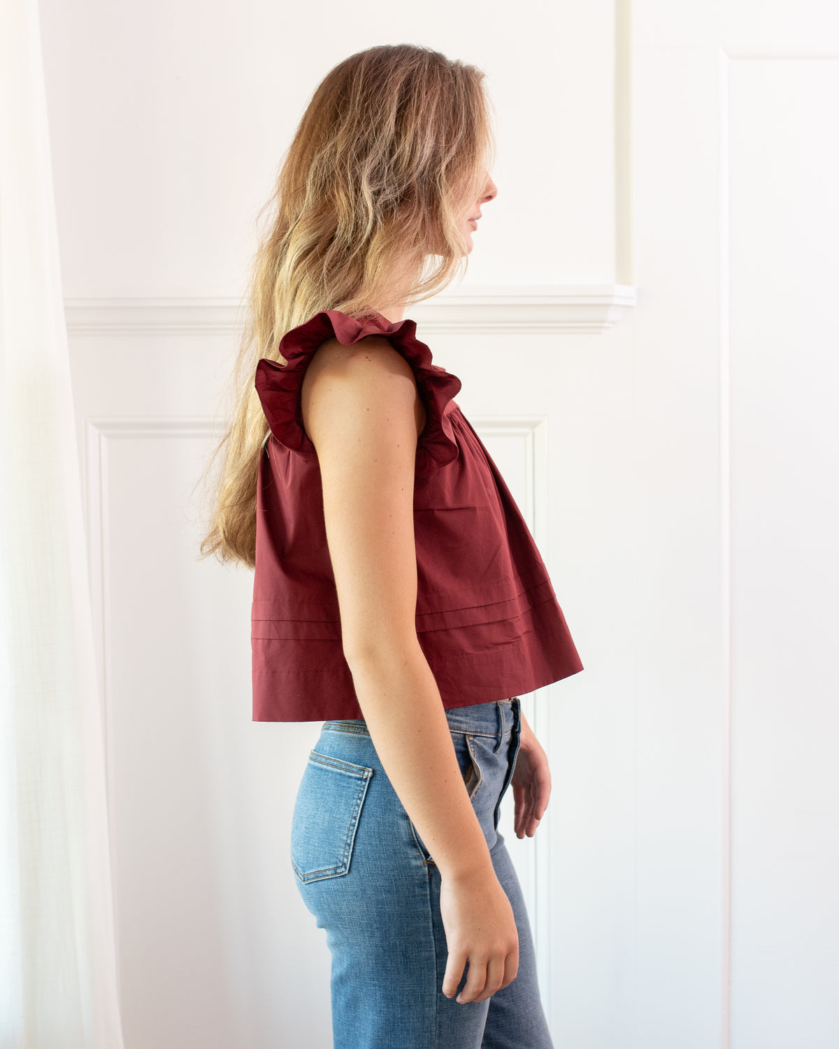 Burgundy cotton flutter sleeve top with pintuck hem worn with jeans, side view.
