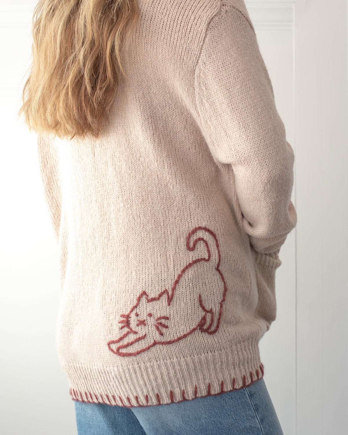 Blush knit cardigan with cat embroidery on the lower back worn by a model, a feminine fall layer, back view.