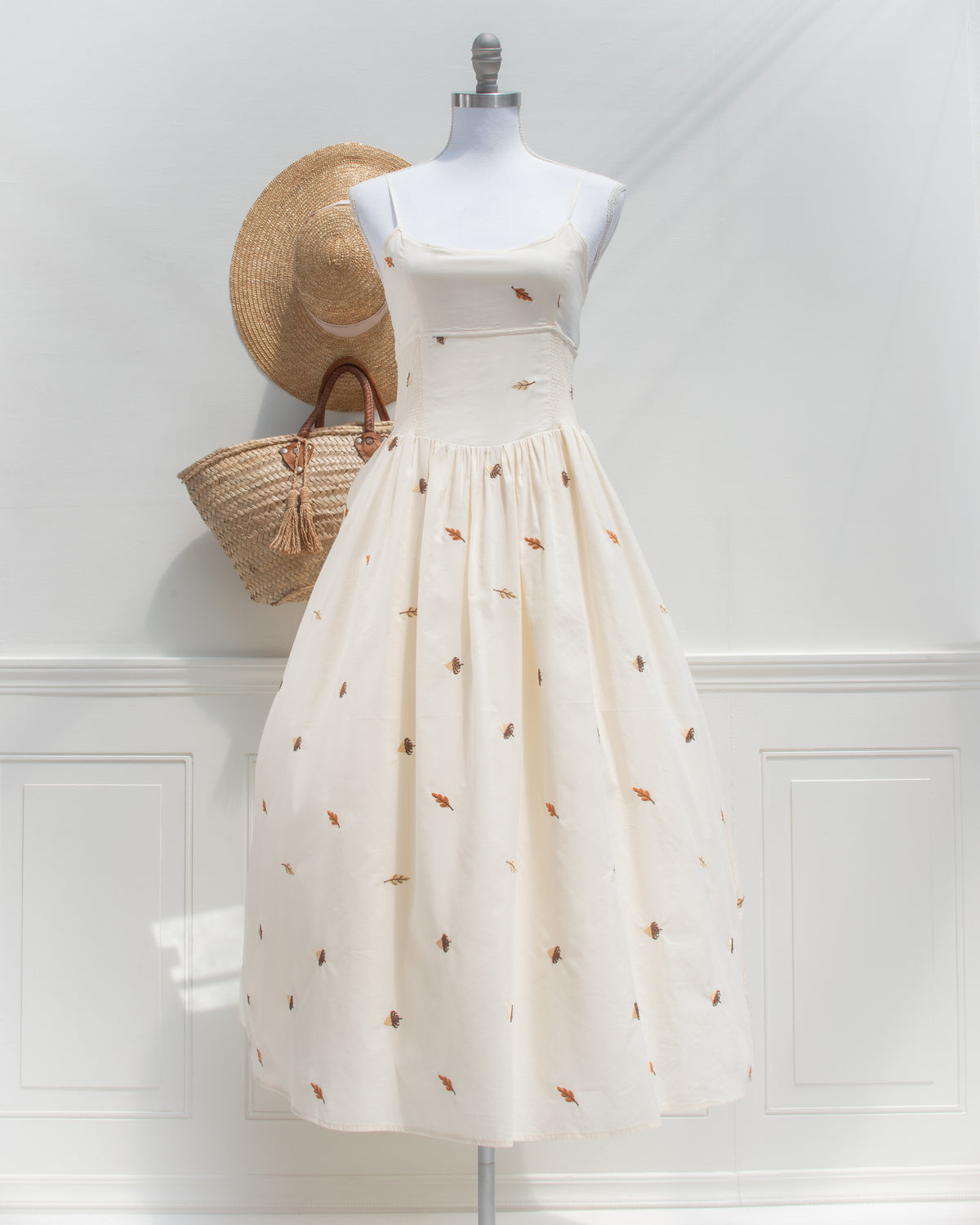 Front view of a cream cotton midi dress with spaghetti straps, smocked waist panels, and embroidered falling leaves and acorns in rust, brown, and gold.