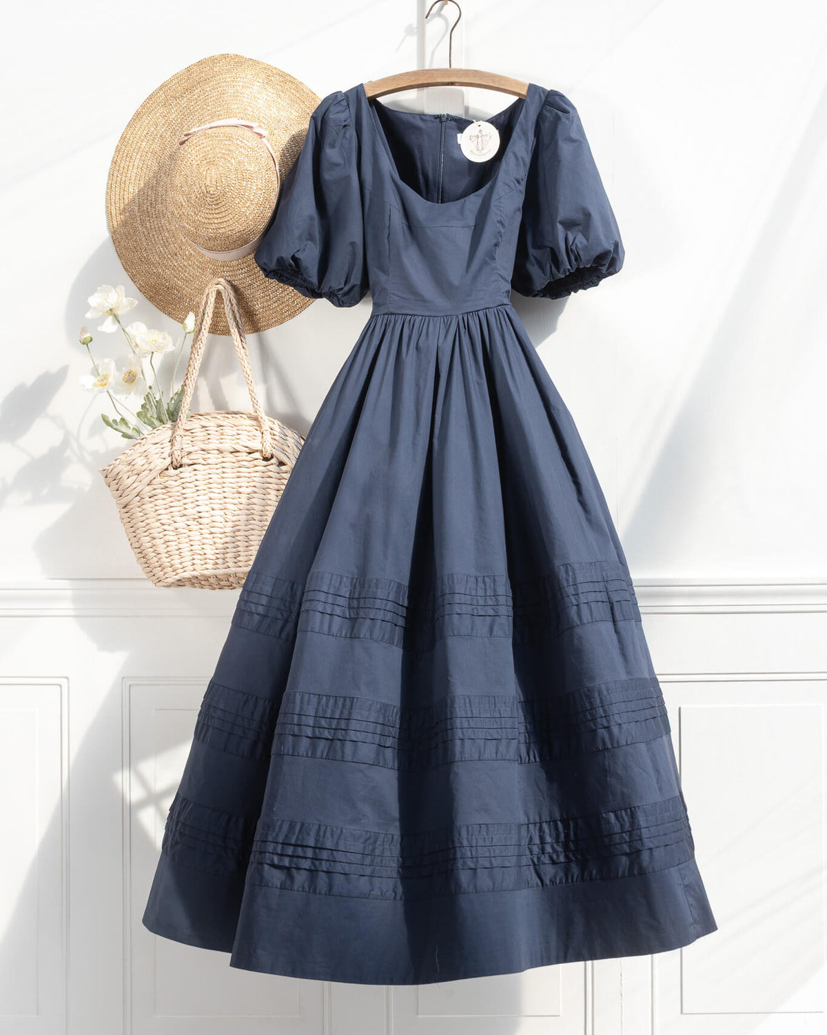 Navy blue dress on a hanger with a straw hat and basket against a white wall.