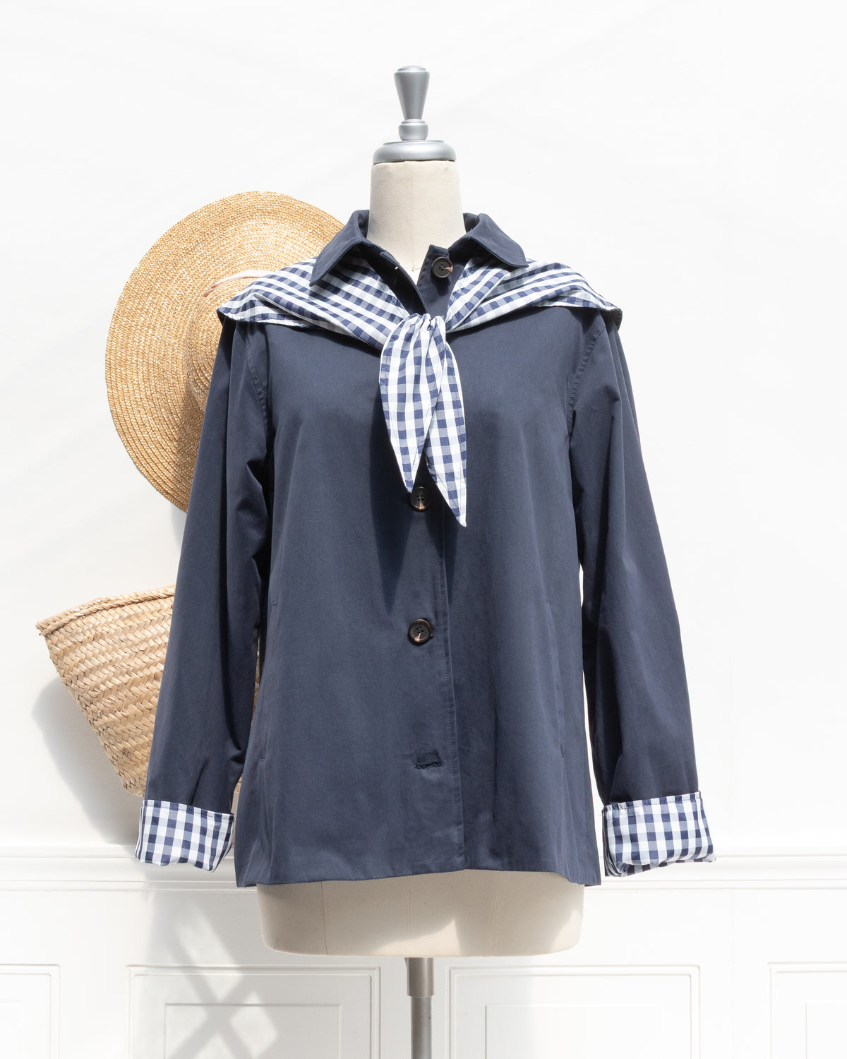 Romantic and feminine clothing, French girl style outerwear. A navy jacket with folded gingham cuffs shown on a mannequin with scarf detail tied at the front, front view. Amantine.