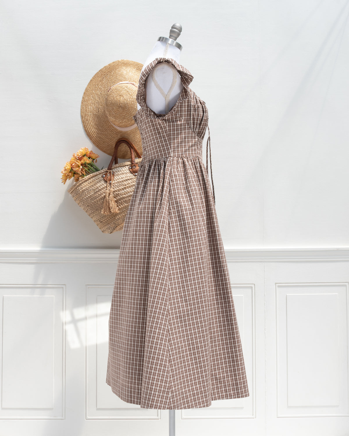 A french style romantic brown and beige checkered midi dress displayed on a mannequin, styled with a straw hat and a woven straw bag against a white paneled wall. side view. 
