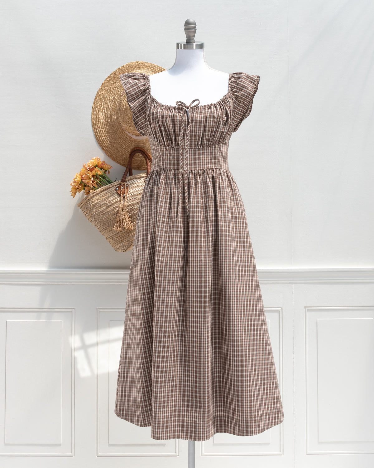 A french style romantic brown and beige checkered midi dress displayed on a mannequin, styled with a straw hat and a woven straw bag against a white paneled wall. front view. 