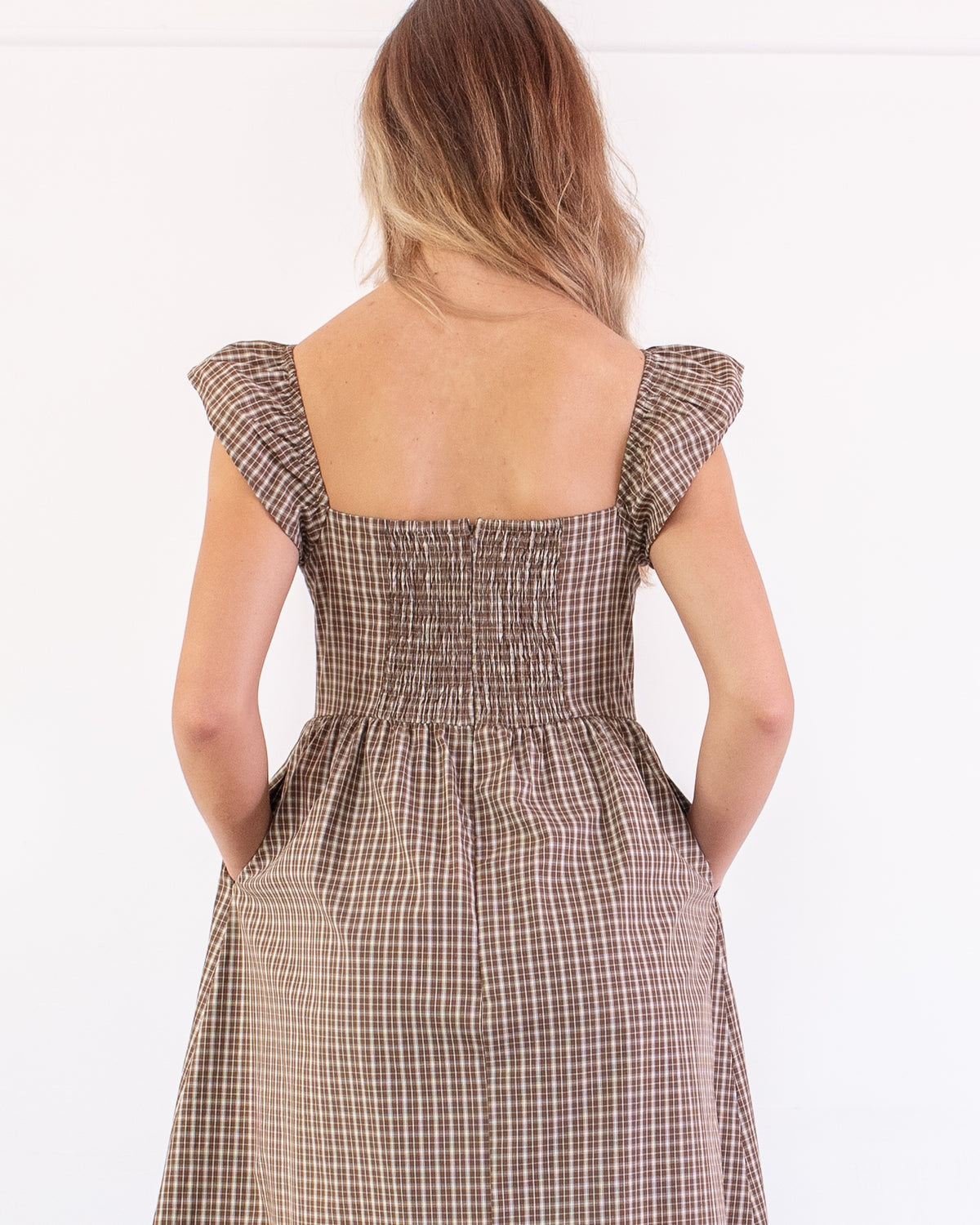 A french style romantic brown and beige checkered midi dress displayed on a model, styled with a straw hat and a woven straw bag against a white paneled wall. back view. 