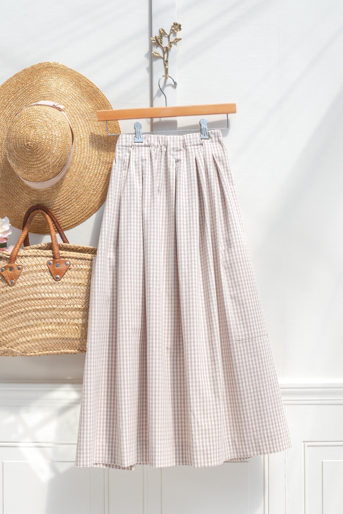 A pleated A-line midi skirt in a beige and white and beige gingham pattern, hanging on a wooden hanger against a white paneled wall. A straw hat and a woven straw tote bag are styled on the left side of the frame. French girl style. 