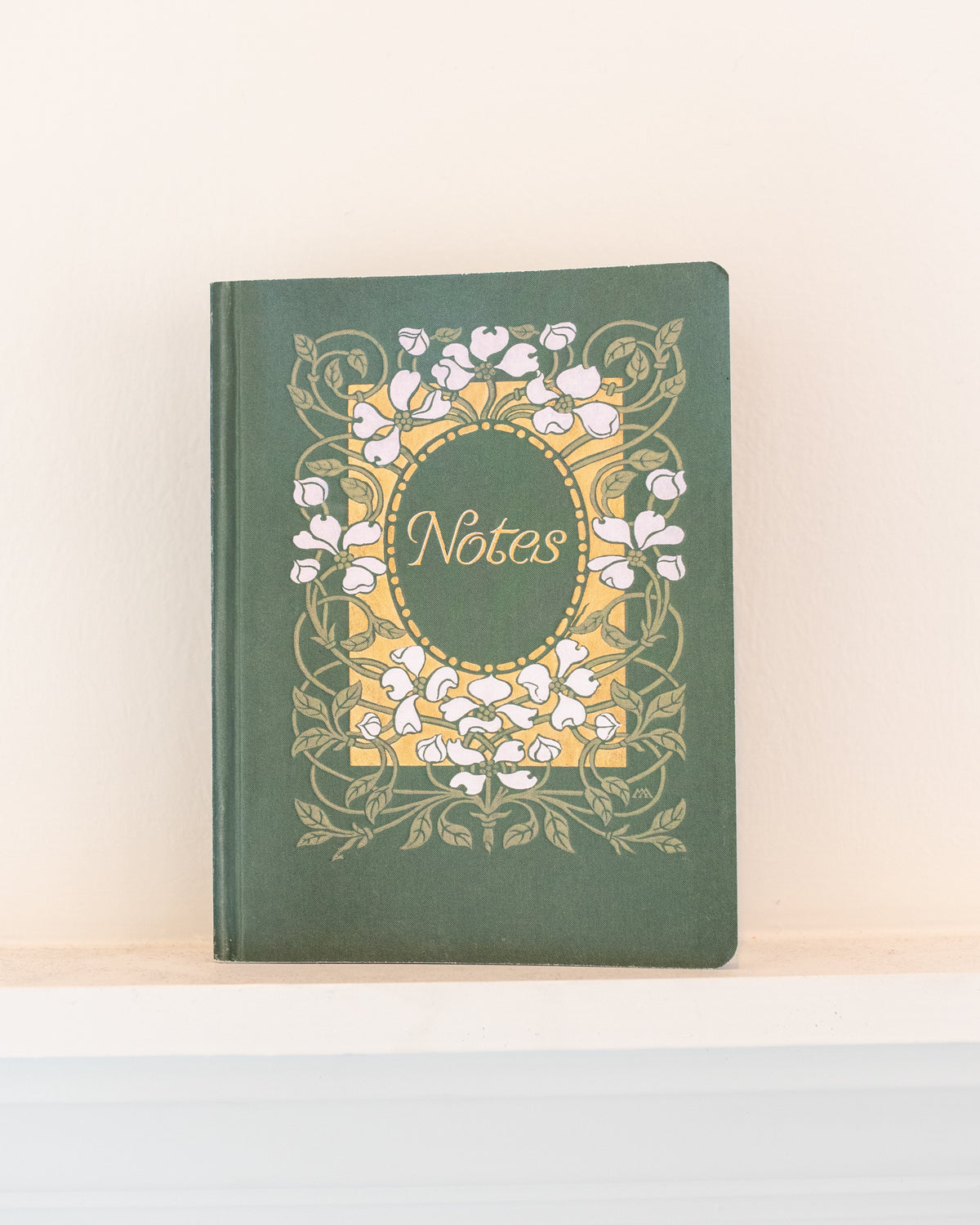 Green vintage notebook with white dogwood blossoms and gold lettering that reads Notes, resting on a shelf, front view.