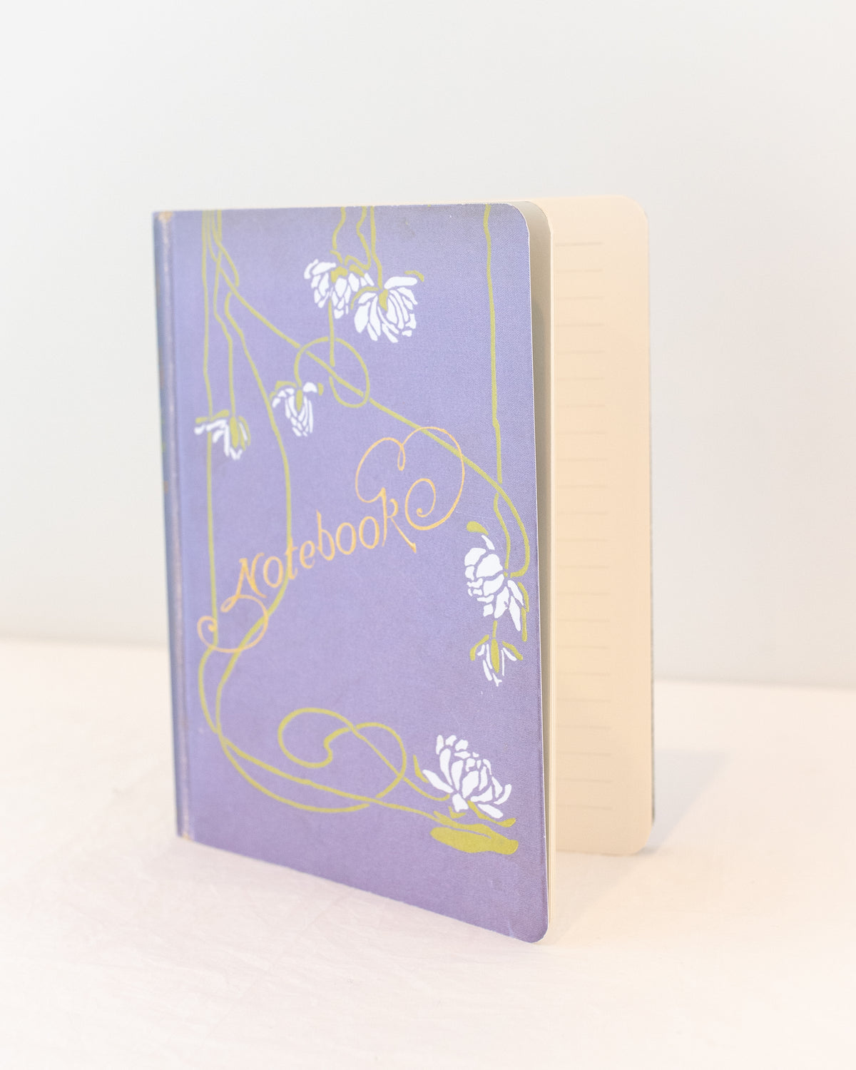 Lavender vintage notebook with white blossoms and gold script lettering, slightly open to lined pages, three quarter view.
