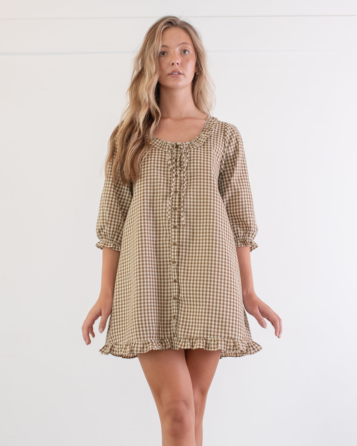 Brown gingham mini dress with tie neckline and button front worn by a model, a classic feminine look, front view.